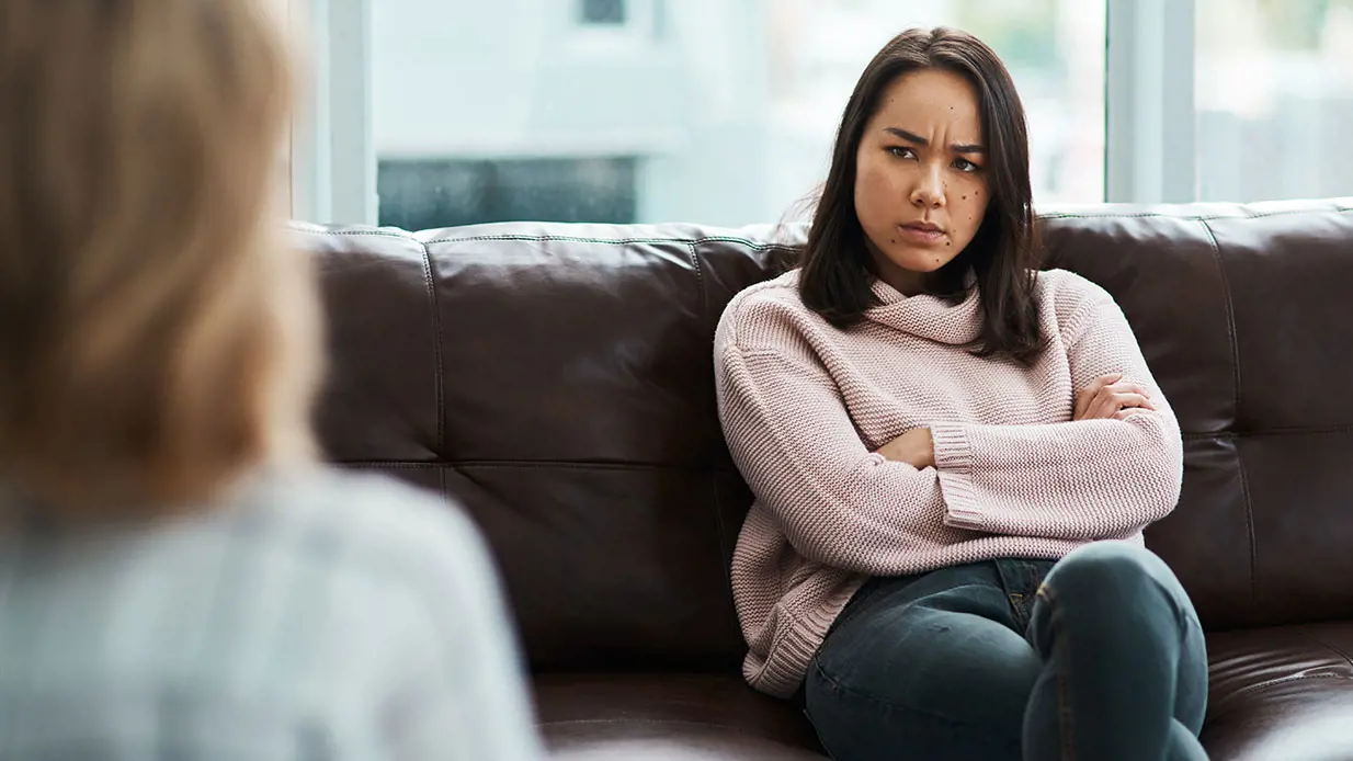 An upset woman sits on a couch listening to someone talk. 