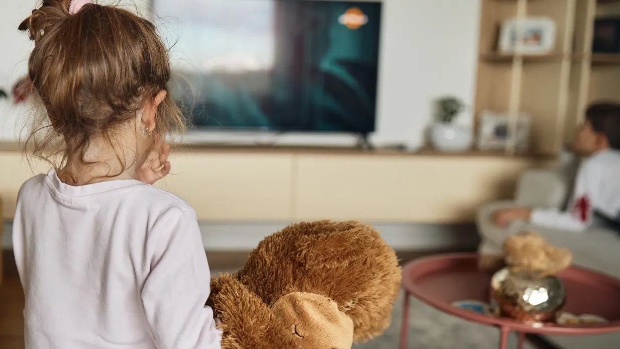 A young girl with a teddy bear stands in front of a television, radiating innocence and joy.