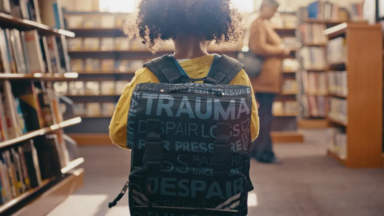 A child wearing a backpack with words like "trauma" and "despair" stands in a library aisle, with an adult browsing shelves in the background.