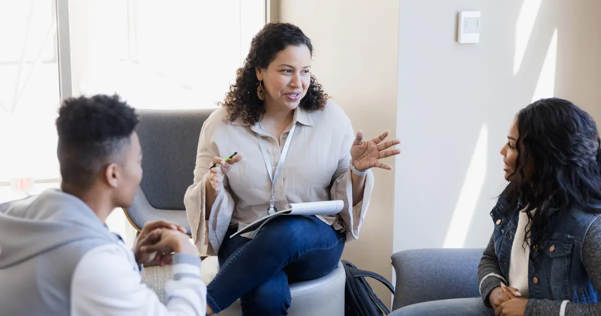 A woman engages in conversation with two students in a classroom setting, fostering an interactive learning environment.