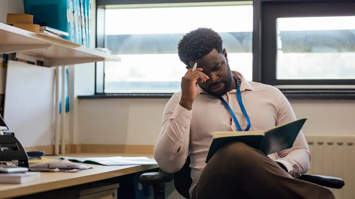 A man sits at a desk, visibly distressed, with his head resting in his hands, conveying a sense of overwhelm.