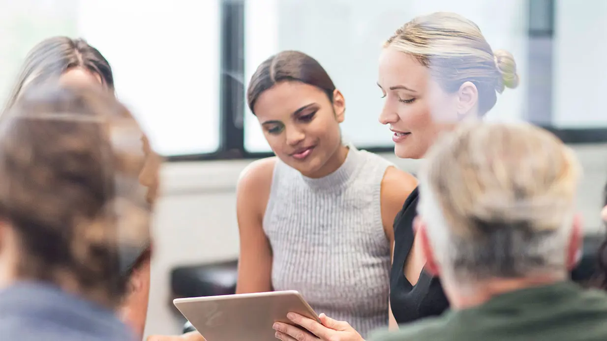 Woman sharing what is on her tablet screen with another woman in a group setting