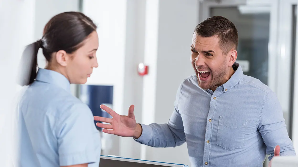 Man in a blue dress shirt yelling at a woman near a receptionist desk