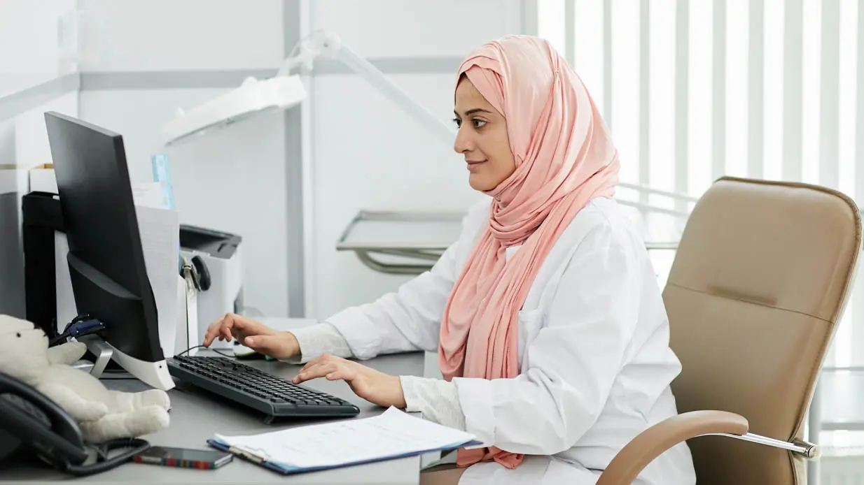 A doctor wearing a hijab working on a computer in an office.