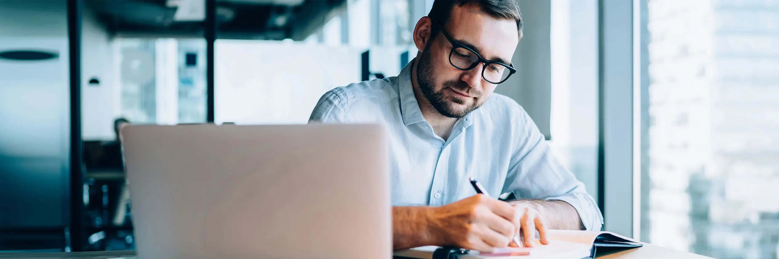 A man jots something down in a notebook while also working on a laptop.
