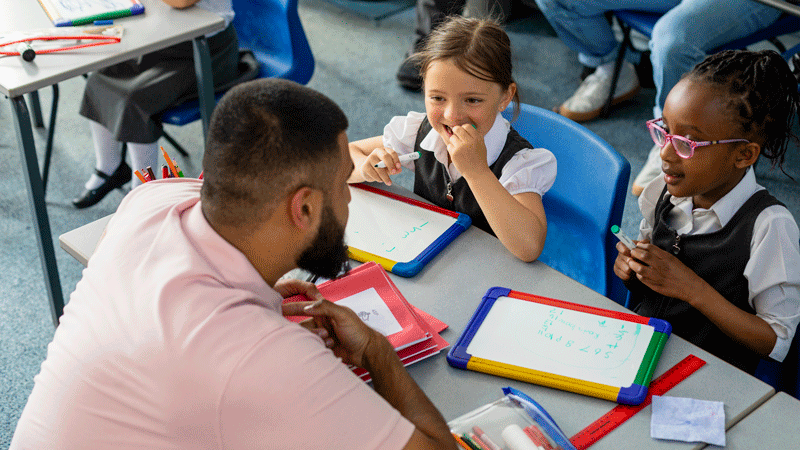 rimary school girls in uniform interacting with their teacher in a classroom setting.