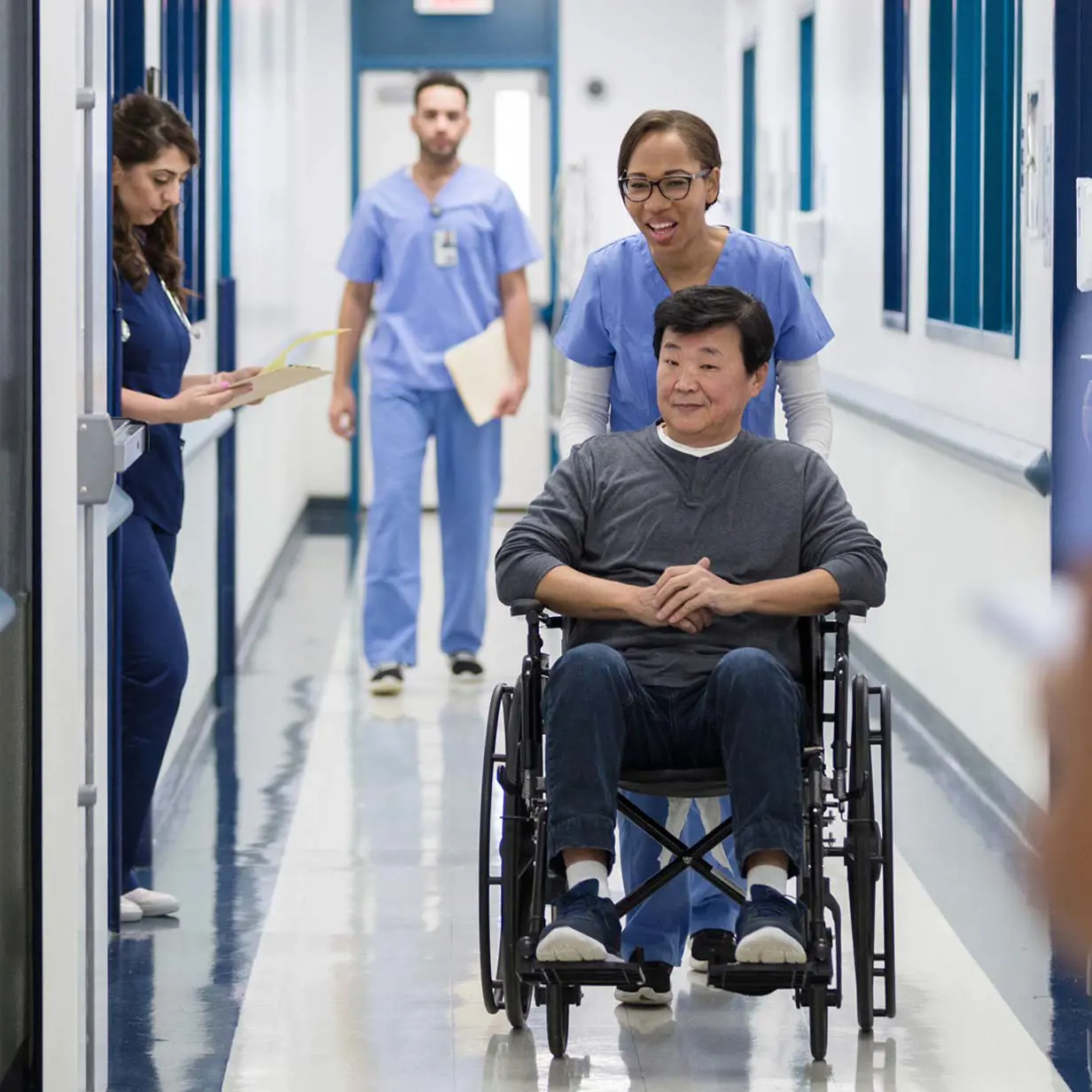 Asian man in a wheelchair being pushed by a nurse in a hallway at a dementia care facility.