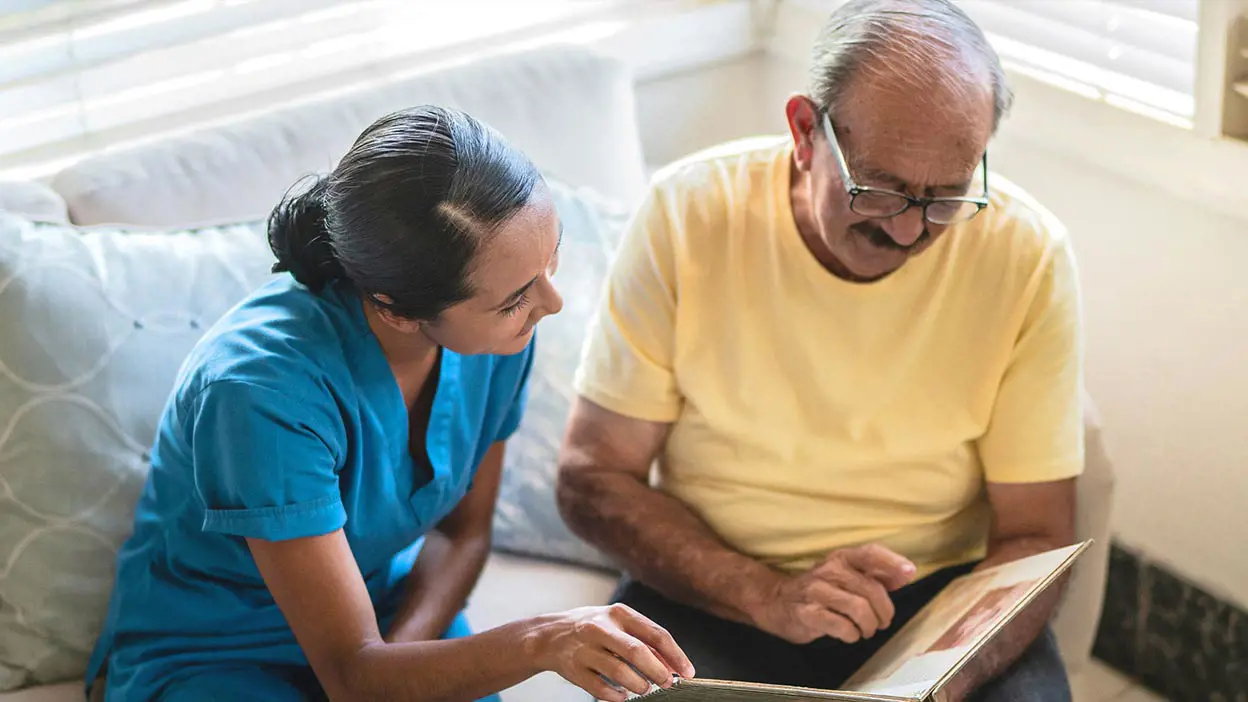 Female nurse in scrubs sitting on a couch with patient looking at photo album.
