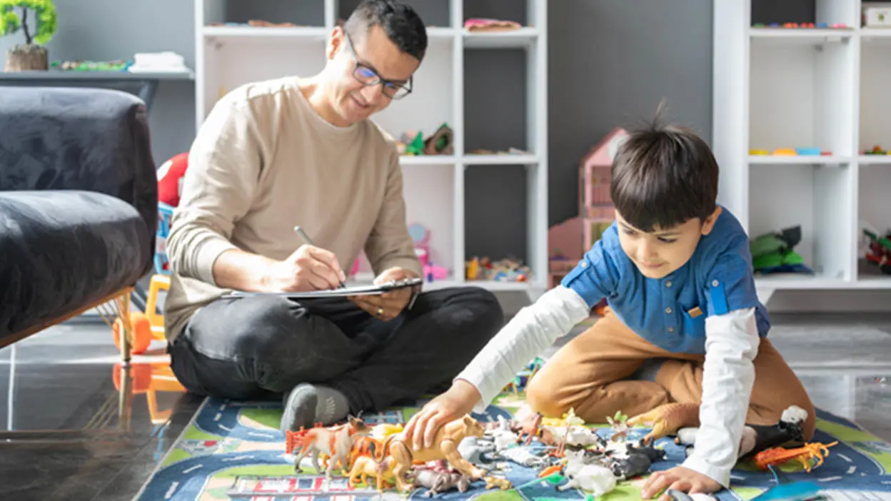 A child plays with toys as an adult looks on taking notes.