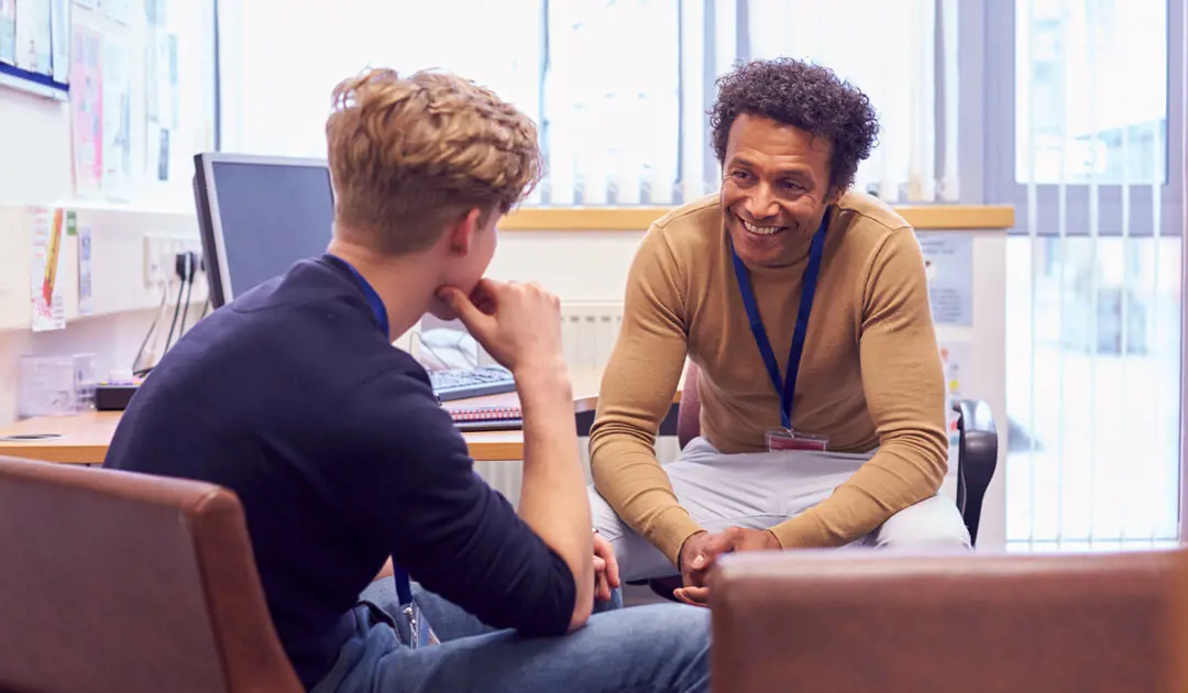 A smiling man talking to a student in his office.